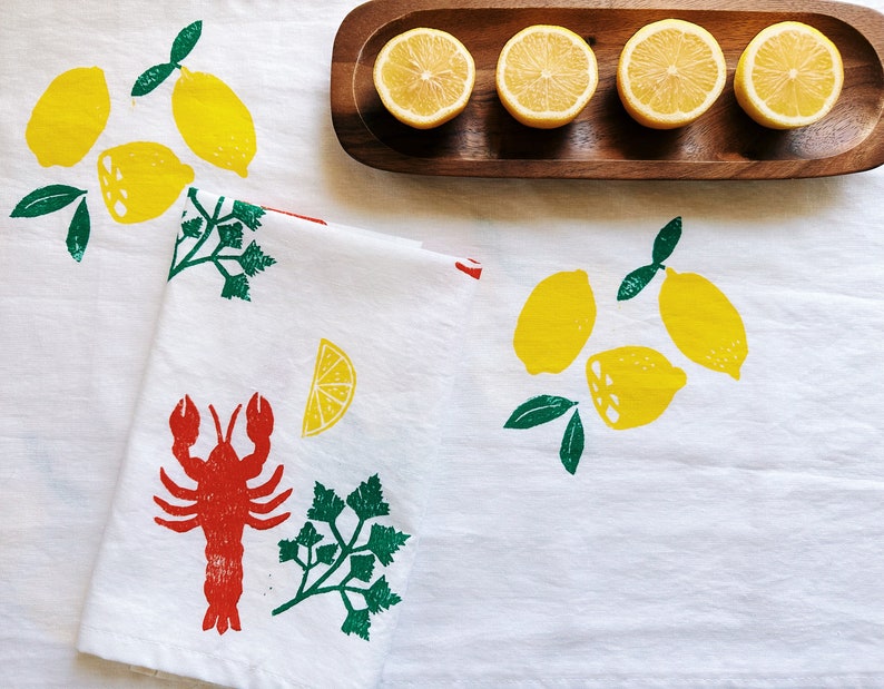 Puede incluir: Un pa&ntilde;o de cocina blanco con un langostino rojo, perejil verde y limones amarillos impresos en &eacute;l. El pa&ntilde;o de cocina est&aacute; doblado por la mitad y est&aacute; sobre un mantel blanco con limones en una bandeja de madera en el fondo.
