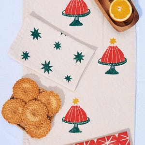 May include: A cream-colored linen table runner and matching napkins featuring block-printed designs of red gelatin molds on green stands topped with yellow stars, green stars, and red circular patterns with white starburst centers.