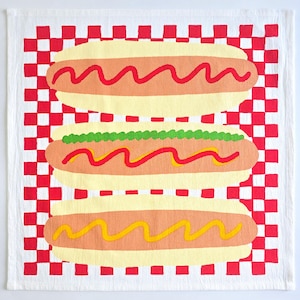 May include: White kitchen towel with a red and white checkered border. The center features three illustrated hot dogs with yellow buns, orange hot dogs, and toppings of ketchup, mustard, and relish.