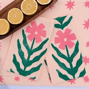 以下が含まれることがあります： ピンクの花柄のキッチンタオル2枚。緑の葉とピンクの花のデザイン。タオルはピンクの星模様のピンク色の表面にあります。木製のトレイにはスライスしたレモンが入っています。
