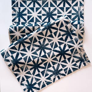May include: White and navy blue geometric patterned fabric. The fabric features a repeating starburst design. The fabric is folded and laid flat on a white surface. The fabric is likely used for home decor or crafting.