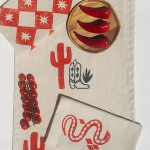 May include: A set of two beige linen kitchen towels with red and black printed designs. One towel features a red checkered pattern with white stars, while the other has a red cactus, boot, and snake design.