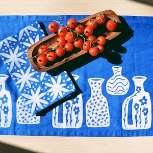 May include: Blue linen tea towel with a white floral pattern and a row of white vases with floral designs. A wooden bowl filled with red tomatoes is on top of the towel.
