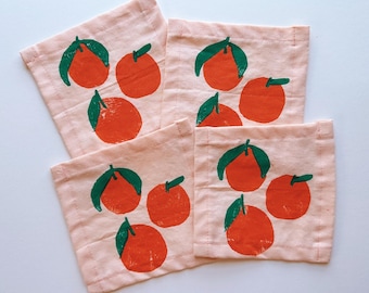 May include: Four square, light pink fabric coasters. Each coaster features a hand-painted design of oranges with green leaves. The oranges are a vibrant orange color. The coasters are arranged on a white surface.