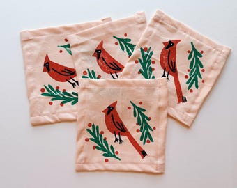 May include: Four square, light pink fabric coasters. Each coaster features a red cardinal bird design with green leaves and red berries. The coasters are likely made of cotton or a similar fabric.