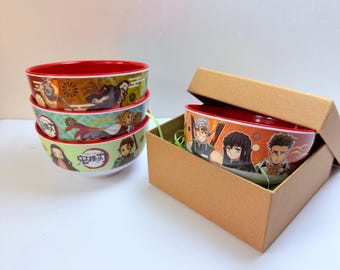 Puede incluir: Un juego de cuatro cuencos de cerámica con coloridas ilustraciones de personajes de anime y texto en japonés. Tres cuencos están apilados, mientras que uno descansa dentro de una caja de regalo cuadrada de cartón. Cada cuenco tiene un interior rojo.