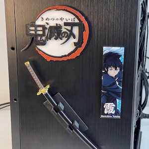 May include: Black computer case with a decorative sword and anime-themed decals. The sword has a gold hilt and a black blade. The decals feature Japanese text and anime characters. The case has a blue light at the top.
