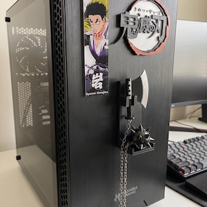 May include: Black computer tower with a tempered glass side panel. The tower has a decorative anime-themed magnet and a hanging weapon charm. The text "Hashira Gaming" is visible on the front.