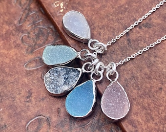 May include: A silver necklace with five teardrop-shaped pendants. Each pendant is made of a different color of crystal: lavender, light blue, gray, blue, and pink.