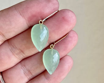 Puede incluir: Un par de aretes delicados con piedras preciosas de color verde claro en forma de lágrima con acabado facetado. Los aretes están suspendidos de ganchos dorados.