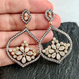 May include: A pair of silver and gold dangle earrings with a floral design. The earrings are adorned with clear and white gemstones.