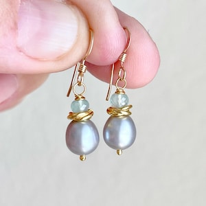 May include: Gold-tone earrings featuring gray teardrop-shaped pearls, light blue beads, and gold wire accents. The earrings have a simple hook design. These earrings are a delicate and elegant accessory.