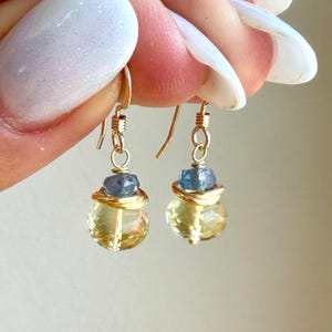 May include: A pair of drop earrings featuring faceted yellow briolette gemstones topped with small blue sapphire beads and gold wire accents. The earrings hang from gold-toned ear wires, showcasing a clear, reflective quality and a dangling design.