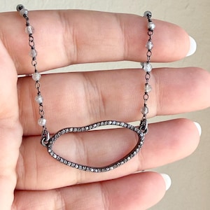 May include: A delicate necklace featuring a dark metal chain and a pendant in an oval shape. The pendant is outlined with small, sparkling stones. The chain also has small, round stones spaced along its length. The necklace is held against a light background.