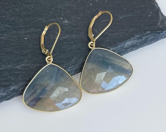 May include: A pair of gold-colored earrings with triangular, faceted, blue-gray gemstones. The earrings have a lever-back closure and a gold-colored frame around the gemstones. The gemstones have a subtle, iridescent quality, reflecting light. The earrings are displayed on a dark, textured surface.