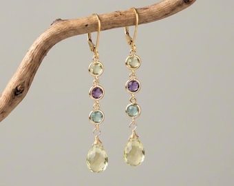 Pode incluir: Um par de brincos pendentes de ouro com três pequenas pedras preciosas circulares em verde claro, roxo e azul, finalizados com uma pedra maior facetada em forma de gota na cor amarelo pálido. Os brincos possuem fechos de alavanca em ouro.