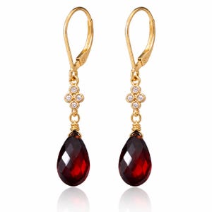 May include: A pair of gold-coloured drop earrings. Each earring features a teardrop-shaped, faceted, deep red gemstone suspended from a gold-coloured chain with a four-petal design set with small, clear gemstones. The earrings have a lever-back closure.