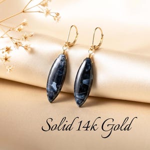 May include: A pair of drop earrings featuring elongated, navy blue and white marbled stone pendants suspended from solid 14k gold leverback hooks. The text Solid 14k Gold is printed below the earrings.