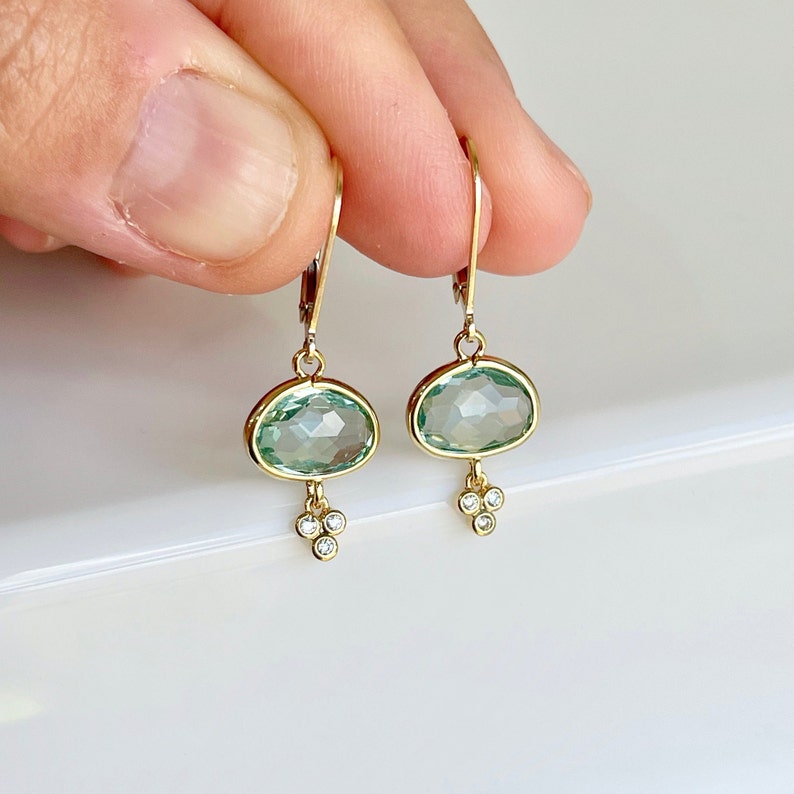 K&ouml;nnte beinhalten: Goldene Ohrh&auml;nger mit einem hellgr&uuml;nen ovalen Edelstein und drei kleinen Diamanten, die unten h&auml;ngen.