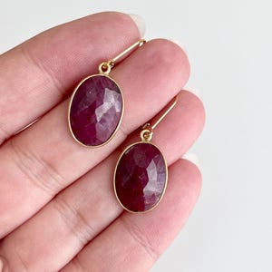May include: A pair of gold earrings with oval-shaped ruby gemstones. The earrings have a simple, elegant design.