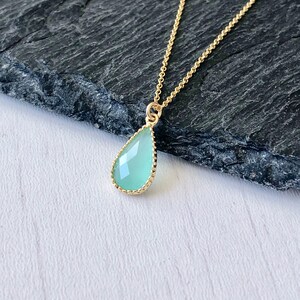 Puede incluir: Un delicado collar de cadena dorada con un colgante en forma de l&aacute;grima de color verde claro. El colgante tiene un borde de cuentas doradas.