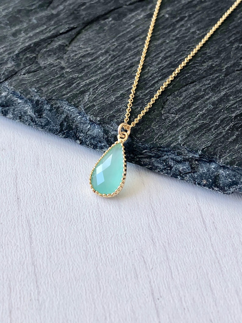 Puede incluir: Un delicado collar de cadena dorada con un colgante en forma de l&aacute;grima de color verde claro. El colgante tiene un borde de cuentas doradas.