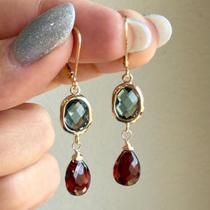 May include: Gold-toned drop earrings featuring a teal-colored, oval-cut gemstone at the top and a teardrop-shaped, deep red gemstone at the bottom. The earrings are suspended from a gold hook.