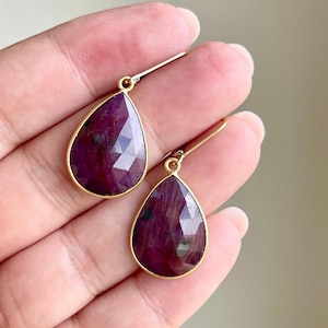 May include: A pair of teardrop-shaped earrings with faceted, deep red gemstones set in gold-colored metal. The earrings are displayed against a neutral background, showcasing their rich color and elegant design. The gemstones have a polished, reflective surface.