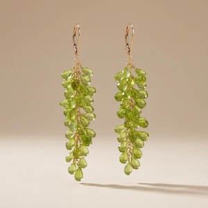 May include: A pair of long dangle earrings featuring numerous small, teardrop-shaped, translucent lime green peridot gemstones clustered together on thin gold-toned wire. Each earring hangs from a gold-toned lever-back closure at the top.