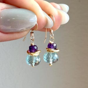 May include: A pair of gold-colored earrings with a three-tiered design. Each earring features a faceted, light blue gemstone at the bottom, a gold-colored ring in the middle, and a dark purple gemstone at the top. The earrings are held up by gold-colored hooks.