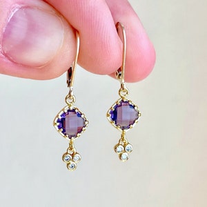 May include: Gold earrings with a square purple gemstone and three small clear gemstones dangling below.