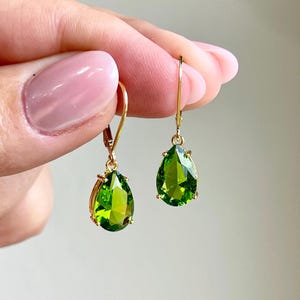 May include: A pair of gold-colored leverback earrings, each featuring a vibrant, pear-shaped, green gemstone. The gemstones are faceted, catching the light and displaying a brilliant sparkle. The earrings are displayed against a soft, neutral background.