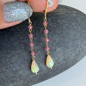 Peut inclure: Boucles d'oreilles pendantes en or avec une chaîne de petites perles roses et une opale en forme de goutte. Les boucles d'oreilles sont suspendues à un crochet en or. Les opales ont un aspect nacré et irisé.