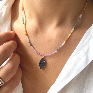 May include: A delicate necklace featuring a multi-colored beaded chain with a pendant. The chain transitions from pink to yellow, gray, and orange. The pendant is a dark, oval-shaped stone with a gold-colored setting. The necklace is worn against a white shirt.