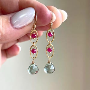 May include: Gold-toned dangle earrings with three oval links, each holding a small, round, red gemstone. The earrings feature a faceted, light green teardrop-shaped gemstone at the bottom. The earrings are held up by a person's fingers.