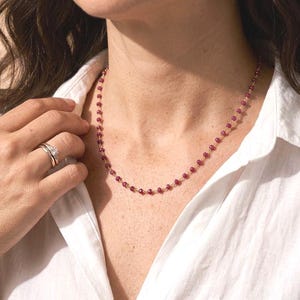 May include: A delicate necklace featuring small, round, red beads strung on a gold chain. The necklace is worn against a white collared shirt. A hand with a ring is visible.