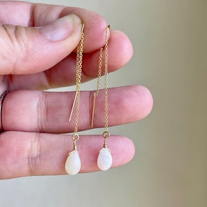 May include: Delicate gold threader earrings with teardrop-shaped, opalescent gemstones. The earrings feature a fine chain and a small gold bar detail. The gemstones are a soft white color.