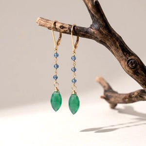 May include: A pair of gold-tone leverback earrings featuring a vertical chain of three small, faceted blue beads. Each earring terminates in a smooth, marquise-shaped green gemstone drop, suspended from the gold wire by a delicate wire-wrapped loop.