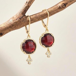 May include: A pair of gold leverback earrings featuring large, round, faceted deep red stones set in gold bezels. Each earring includes a small, triangular cluster of three tiny clear stones dangling from the bottom of the main setting.
