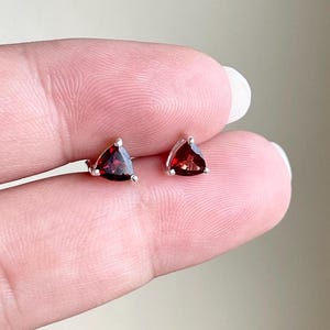 May include: A pair of stud earrings featuring deep red, triangular-cut garnet gemstones set in polished silver metal. Each earring has a three-prong setting that secures the faceted stone, creating a classic and symmetrical appearance for the jewelry.