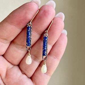 May include: A pair of gold earrings with a teardrop opal dangle. The earrings feature a bar of blue sapphires set in a black metal frame.