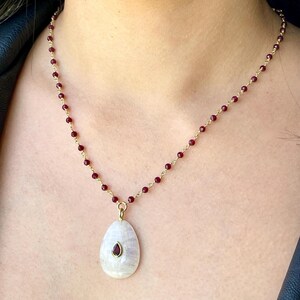 May include: A gold chain necklace with small red beads and a pear-shaped pendant. The pendant is white with a small red gemstone in the center. The necklace is worn around the neck.