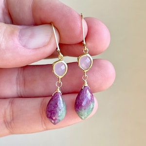 May include: Gold earrings with pink and green gemstone drops. The top of the earrings feature a square-shaped pink gemstone.