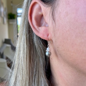 May include: A silver earring with a gray pearl and a small white bead. The earring is hanging from a silver hook. The woman's ear and hair are visible in the image.