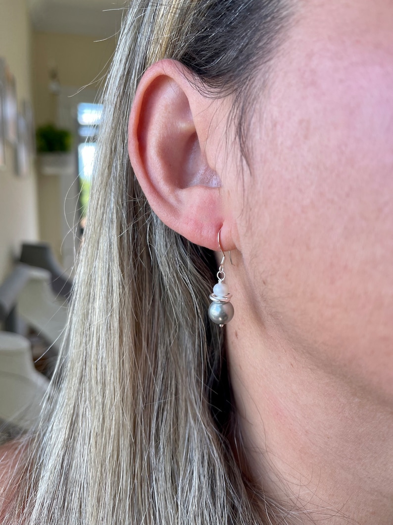 May include: A silver earring with a gray pearl and a small white bead. The earring is hanging from a silver hook. The woman's ear and hair are visible in the image.