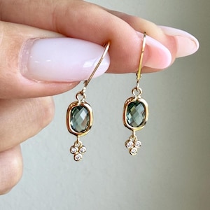 May include: A pair of gold drop earrings featuring faceted oval green gemstones set in gold bezels, with a small cluster of three tiny clear round stones dangling from the base of each oval, suspended from thin gold ear wires.