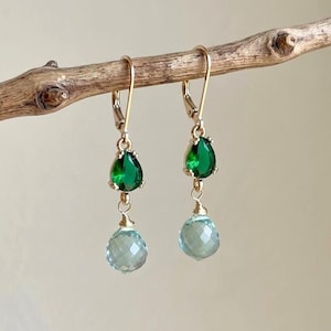 May include: A pair of gold-tone leverback earrings featuring a faceted emerald green teardrop-shaped stone at the top and a round, faceted light blue crystal bead suspended below by a gold-tone wire wrap, hanging from a textured wooden branch.