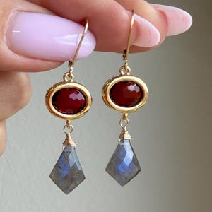 May include: A pair of gold-tone drop earrings featuring an oval-shaped faceted deep red stone set in a gold frame, connected to a dangling kite-shaped faceted grey labradorite stone with blue iridescence, suspended from gold-tone ear wires.