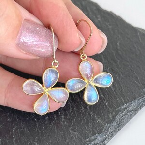 May include: Gold-tone earrings featuring four-petal flower designs. Each petal is a pearlescent, iridescent stone with blue and white hues. The earrings are held up by a gold hook.