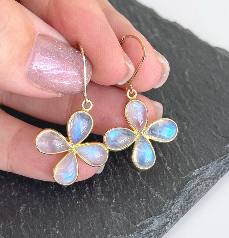 May include: Gold-tone earrings featuring four-petal flower designs. Each petal is a pearlescent, iridescent stone with blue and white hues. The earrings are held up by a gold hook.
